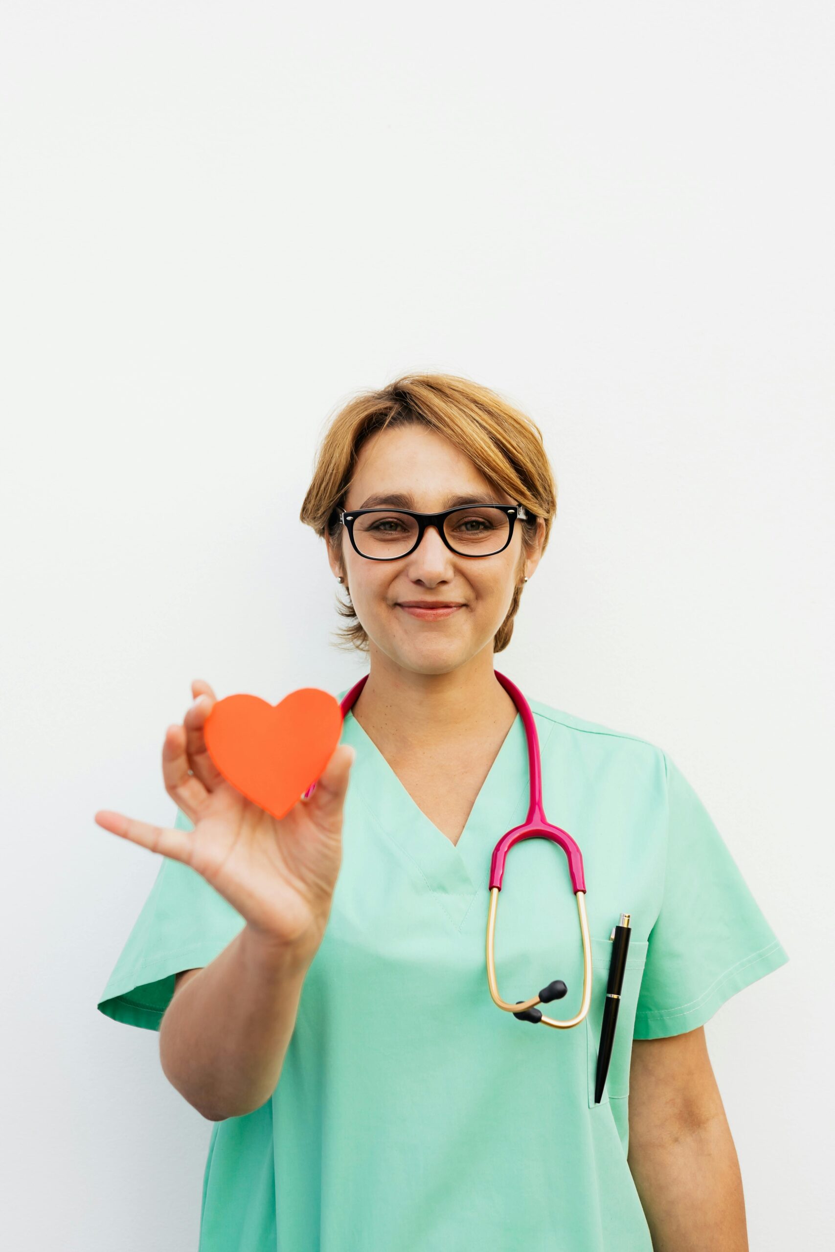 Smiling healthcare professional holding a heart symbol, standing confidently with a white background.